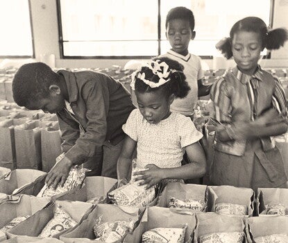 A black-and-white photograph showing children preparing bags of food to be distributed through the Panther Free Food Program at the Black Panther Community Survival Conference in Oakland, California. Four Black children, two boys and two girls, are placing bags of puffed wheat cereal into brown paper grocery bags that have been organized in rows.