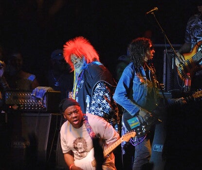 George Clinton, a Black man, wearing a bright red wig, dark blue vest, and multicolored shirt with skulls and crossbones, playing the keyboard as other musicians surround him.
