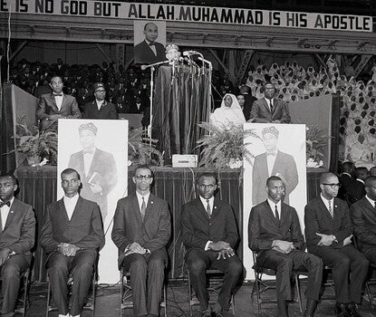 Elijah Muhammad, a Black man wearing a beret, barely visible above a lectern and a bank of microphones. He is flanked on his stage by two Black men on his right (one in a dark suit, white shirt, and dark bowtie, and another in a Fruit of Islam uniform of beret, dark jacket, and white shirt) and a Black woman (in a white robe and headwear) and Black man (in a dark suit, white shirt, and dark necktie), and behind him are several Black men in dark suits and ties and Black women in white robes and headwear.