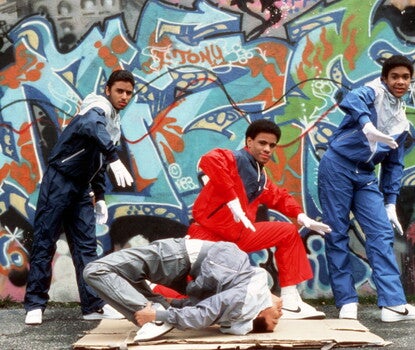 Three young Black men wearing tracksuits and white gloves dancing as another young Black man inverts his body on a piece of cardboard. Behind them is a graffitied wall.
