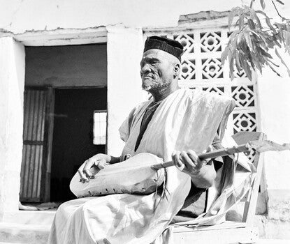 A black-and-white photograph of an elderly Black man in a white robe and black kufi hat sitting with eyes closed, plucking a guitar made of a calabash squash.