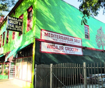 A color photograph of a corner store with a two-toned green wall with two signs, one reading "Wadajir Grocery, Finest Halal Meats" and the other "Mediterranean Deli, Best Gyros in Town"