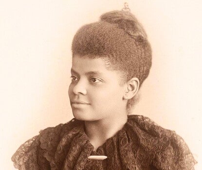 The image is a sepia-toned portrait of Ida B. Wells, the African American journalist, activist, and anti-lynching crusader. She is shown from the chest up, wearing a high-collared, intricately embroidered lace dress with ruffled details. Her hair is styled in an elegant updo, and she has a composed, determined expression while gazing slightly to the side. The soft lighting and classic photographic style suggest it was taken in the late 19th or early 20th century.