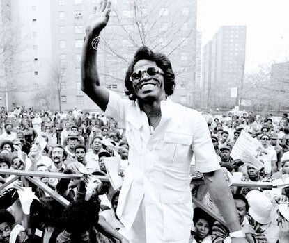 A black-and-white photograph of James Brown, a Black man wearing a white shirt, sunglasses, a metal bracelet, and wristwatch waving as fans wave pieces of paper and look on in the distance.