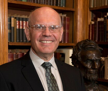 A bald white man wearing rectangular glasses and a suit smiles at the camera. He is standing in front of a bust of Lincoln and a bookshelf.