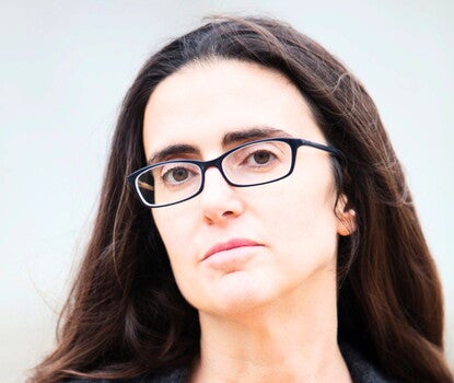Headshot of a white woman with long brown hair and black rectangular glasses, wearing a red shirt and gray sweater, looking directly at the camera. 