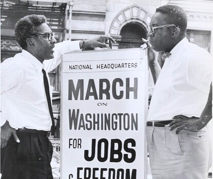 Two Black men stand talking on either side of a large sign reading "National Headquarters/March on Washington for Jobs and Freedom/Wed. Aug. 28". 