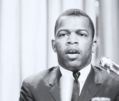 John Lewis, a Black man, wearing a dark jacket and tie and white shirt, speaking in front of two microphones on a table.