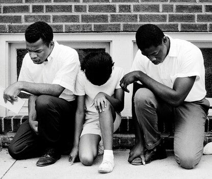 From left to right: a Black woman wearing a white vest and plaid pants kneeling with head raised; John Lewis, a Black man wearing a white button-down shirt, black pants, and black shoes, kneeling with head bowed while holding a notepad in his left hand; a Black girl wearing a white shirt, shorts, and shoes, kneeling with head bowed; and a Black man wearing a white button-down shirt, black pants, and black shoes, kneeling with head bowed. All are in front of a brick building with ground-floor windows.