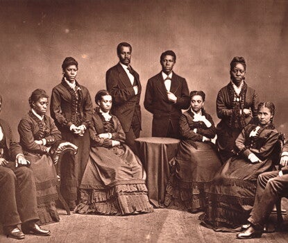 Ten Black men and women sitting and standing while wearing dark suits or dresses. A small table with a dark tablecloth sits in the middle background.