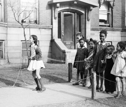 A black-and-white photograph of a young Black girl wearing a white dress, dark sweater, and dark socks and shoes, jumping rope as other Black children look on.