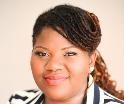 A Black woman with reddish-brown hair and hands together at the front of her body, wearing a white shirt, black-and-white striped blazer, and earrings.