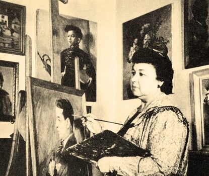 Laura Wheeler Waring, a Black woman, wearing an artist's smock and painting a portrait of a man with her right hand while holding a palette in her left. On the walls behind her are other portraits and artwork.