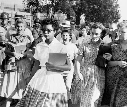A Black girl wearing sunglasses, a white blouse, and a white and black/white checkered skirt, holds a book in the crook of her left arm while walking to school. A White girl in the background screams at the Black girl as other White boys and girls, and men wearing helmets, look on.