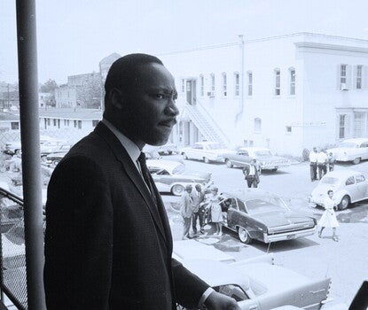 Martin Luther King Jr. in a dark suit and tie and white shirt, looking out from a balcony at the street below.