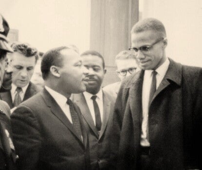 Malcolm X and Martin Luther King Jr. smiling and talking with each other while a police escort stands near. 