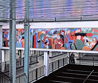 A photograph with impressionist, multicolored mosaic mural of people on a subway train in the background, with beams holding up a subway station in the foreground of the photograph.