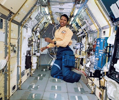 Mae Jemison, a Black woman, wearing an astronaut uniform (yellow shirt with mission patch, blue cargo pants, grey slippers) aboard the Space Shuttle. She is floating in zero gravity, surrounded by experimental stations, with a video camera floating to her left.