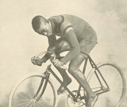 A black-and-white photograph of Major Taylor, a Black man, in a dark striped shirt and shorts atop a racing bicycle.