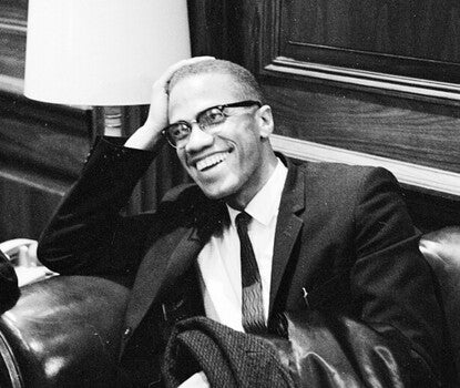 Malcolm X, a bespectacled Black man, wearing a dark suit, dark patterned tie, and white shirt, with a large ring on his left finger. He is sitting in an upholstered chair or sofa and smiling with his head in his right hand (leaning on the armrest of the chair or sofa).