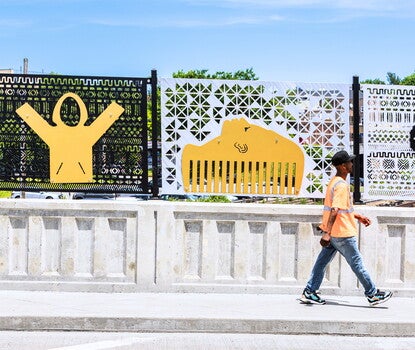 A Black man wearing a black baseball cap, orange shirt, jeans, and sneakers, walking along a bridge with a series of metal panels in the background.