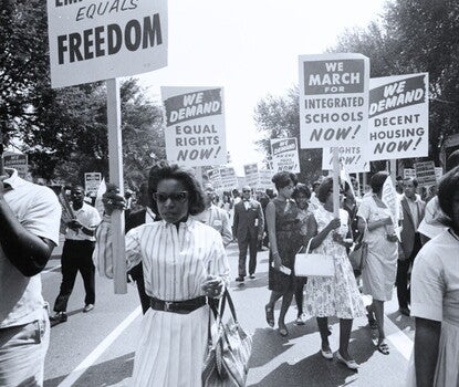 Photograph shows a procession of African Americans, with two women in the foreground, carrying signs for equal rights, integrated schools, decent housing, and an end to bias. 
