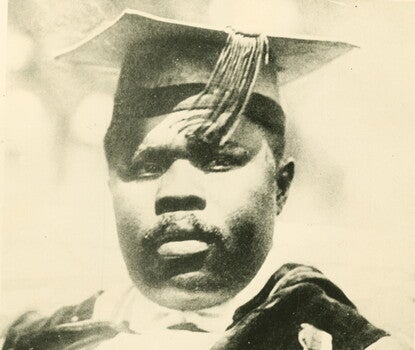 Marcus Garvey, a Black man, wearing a dark jacket and vest, white shirt, and dark tie. He is also wearing a button on his lapel.