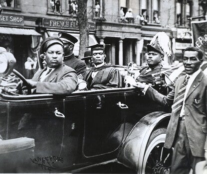 An open-topped car with four passengers in a parade. The driver is a Black man with a dark jacket, white shirt, dark tie, and dark driver's cap. In the back seat, from left to right, is a Black man in a military uniform; a Black man wearing academic regalia; Marcus Garvey, a Black man, wearing a military uniform and holding a sword. Next to the car is a Black man wearing a dark jacket, white shirt, and striped tie. In the background are apartment buildings, where Black people are looking on at the parade.
