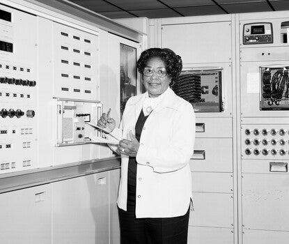 Mary Jackson, a bespectacled Black woman wearing a white lab coat over a dark sweater, white shirt, and brooch, writing on a clipboard as engineering instruments surround her.