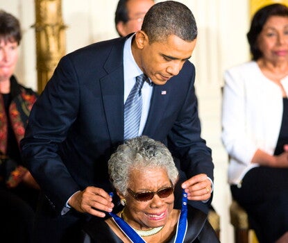 President Barack Obama, standing wearing a black suit, white shirt, and striped gray and white tie, awarding the Presidential Medal of Freedom to Maya Angelou, seated wearing a black dress, sparkly necklace and earrings, and sunglasses, as other honorees (including Sylvia Mendez) sit and look on. 