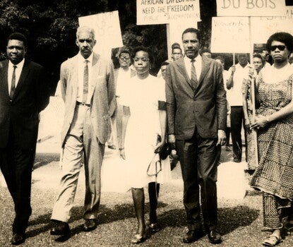 Three Black men in suits and two Black women in dresses protesting, as other people protest in the background. The woman on the right, Maya Angelou, is holding a sign, the bottom of which reads "W.E.B. Du Bois."