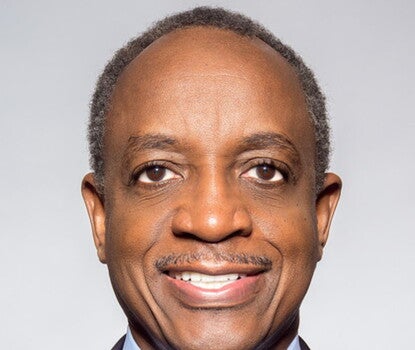 A Black man smiling while wearing a black suit, light blue shirt, and patterned red tie.