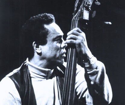 Charles Mingus, a Black man wearing a white or light-colored turtleneck and a dark vest, playing an upright bass.