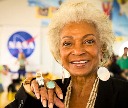 Nichelle Nichols, a Black woman with white hair, wearing a black shirt, beaded necklace, and white round earrings. She is holding a small LEGO character of an astronaut in a white spacewalking suit, next to a large gold and blue ring on her finger.