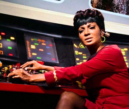 Lieutenant Uhura (Nichelle Nichols), a Black woman, wearing her Starfleet uniform (red dress with a black collar and a wavy gold band on each sleeve), with a communications earpiece in her left ear, along with green hoop earrings. She has her hands on a bank of buttons connected to an array of four light screens.