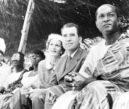 Seated, from left to right, a Black man wearing a white shirt, kente cloth robe, and glasses; a Black woman wearing a white shirt; Pat Nixon, a White woman, wearing a light-colored dress with a white collar and a white hat; Richard Nixon, a White man, wearing a dark suit, white shirt, and dark tie; and K.A. Gbedeman, a Black man, wearing a light-colored shirt and kente cloth robe.
