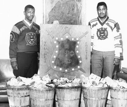Two young Black men in long-sleeved T-shirts emblazoned the Omega Tau Psi crest across the chest, flanking a large crest for the fraternity, in front of five barrels full of canned and packaged food.