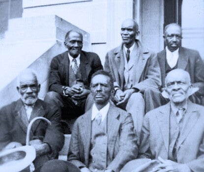 Six older Black men wearing dark suits sitting on steps in two rows of three.