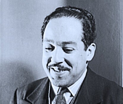 The image is a black-and-white portrait of Langston Hughes, the renowned African American poet, playwright, and leader of the Harlem Renaissance. He is shown from the chest up, wearing a suit with a tie and a sweater vest underneath. He has a warm smile and is looking slightly downward, with soft lighting casting gentle shadows on his face. The background is plain, and the photograph has a classic, vintage feel, likely taken in the mid-20th century.