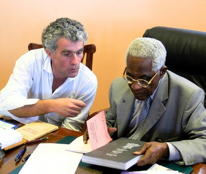 Phillippe Mouillon, a gray-haired White man (wearing a collared white shirt), sitting next to Aimé Césaire, a Black man, wearing a gray jacket, gray shirt, and patterned blue and white tie. Both are sitting in chairs (Mouillon in a wooden chair, and Césaire in a padded office chair upholstered in black fabric), and Césaire is holding a book in each hand.