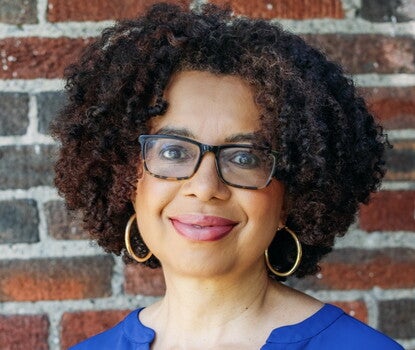 A woman with curly, short, black and auburn hair smiles at the camera in front of a brick background. She wears a blue top, gold hoop earrings, and glasses with black frames. 