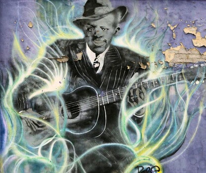 Photograph of a mural of Robert Johnson (a Black man holding a guitar while wearing a dark suit, white shirt, dark tie, and dark hat), painted in black and white and framed by wisps of smoke, on a purple wall. 