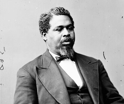 A black-and-white photograph of Robert Smalls, a goateed Black man, seated, wearing a dark jacket, bow tie, and vest (with pocket watch chain), and a white shirt.