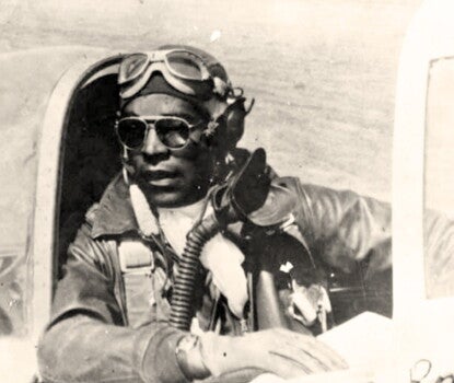 A Black pilot in aviation gear sits in the cockpit of his plane, looking over his shoulder. 