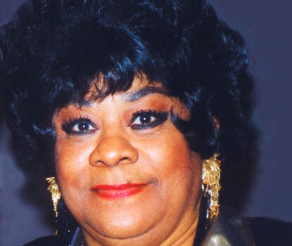 A color headshot of Ruth Brown, a Black woman, wearing a black jacket with a sparkly gold collar, two metallic necklaces, and dangling gold earrings.  