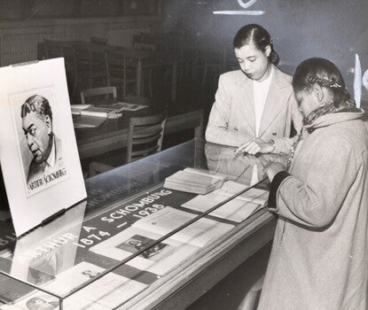Two Black women wearing light-colored coats look into a glass vitrine case with the title "Arthur A Schomburg, 1874–1938." Atop the case is a matted photograph of Arturo [Arthur] Schomburg.
