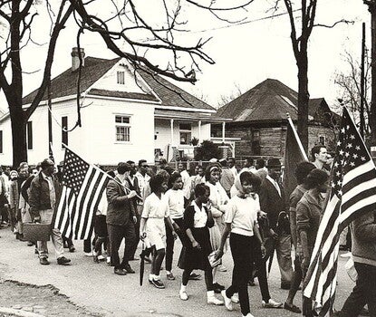 Scan of a black and white photograph of a mix of Black men, women, and children and a few white men and women, some carrying American flags,  walking through a town. 
