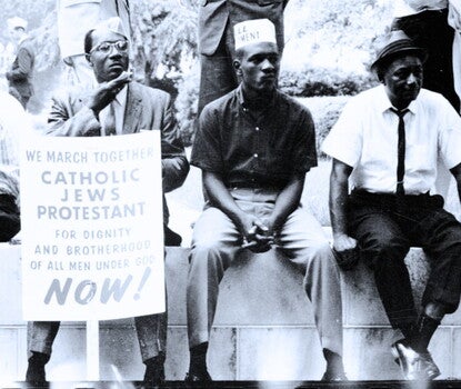 Three Black men sit along a wall. One holds a sign that reads "We march together/ Catholic, Jews, Protestant/ for dignity an brotherhood of all men under God now!" 