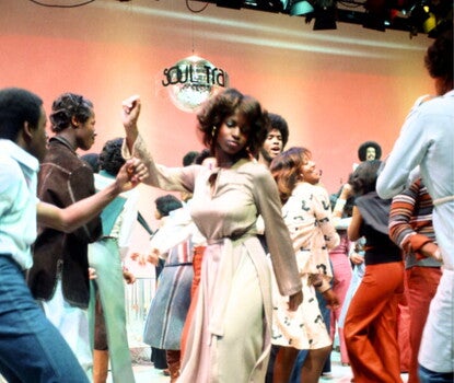 A Black woman wearing a beige jumpsuit with a cinch dancing under a mirrorball reading "Soul Train," as other Black men and women dance around her.