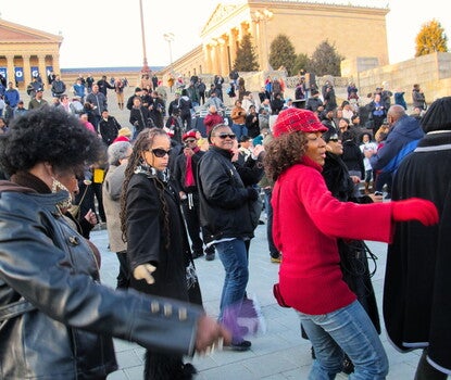 Black women in coats and sweaters dancing as a multiracial crowd looks on from the steps of the Philadelphia Museum of Art (with the Museum's colonnade in the background).